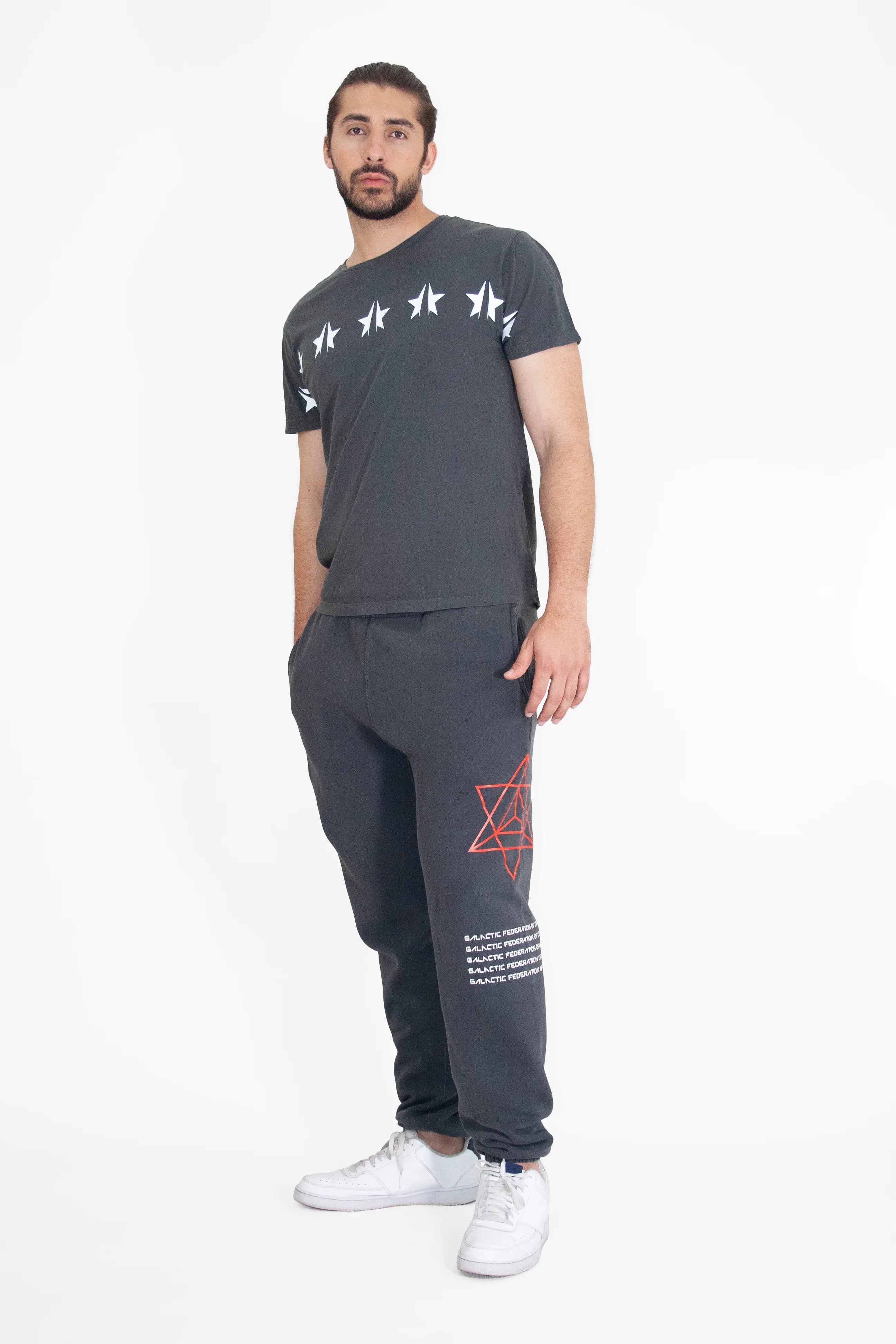 A person wearing a black GFLApparel GFL Stars Tee in Space Glow featuring white star graphics and black sweatpants with red geometric designs stands against a plain white background. The relaxed fit and breathable cotton promise comfort throughout the day.