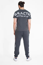 A person with a bun stands facing away, wearing a dark gray GFLApparel GFL Stars Tee in Space Glow with "GALACTIC FEDERATION OF LIGHT" written on the back. The relaxed fit t-shirt, made from breathable cotton, is paired perfectly with matching sweatpants.