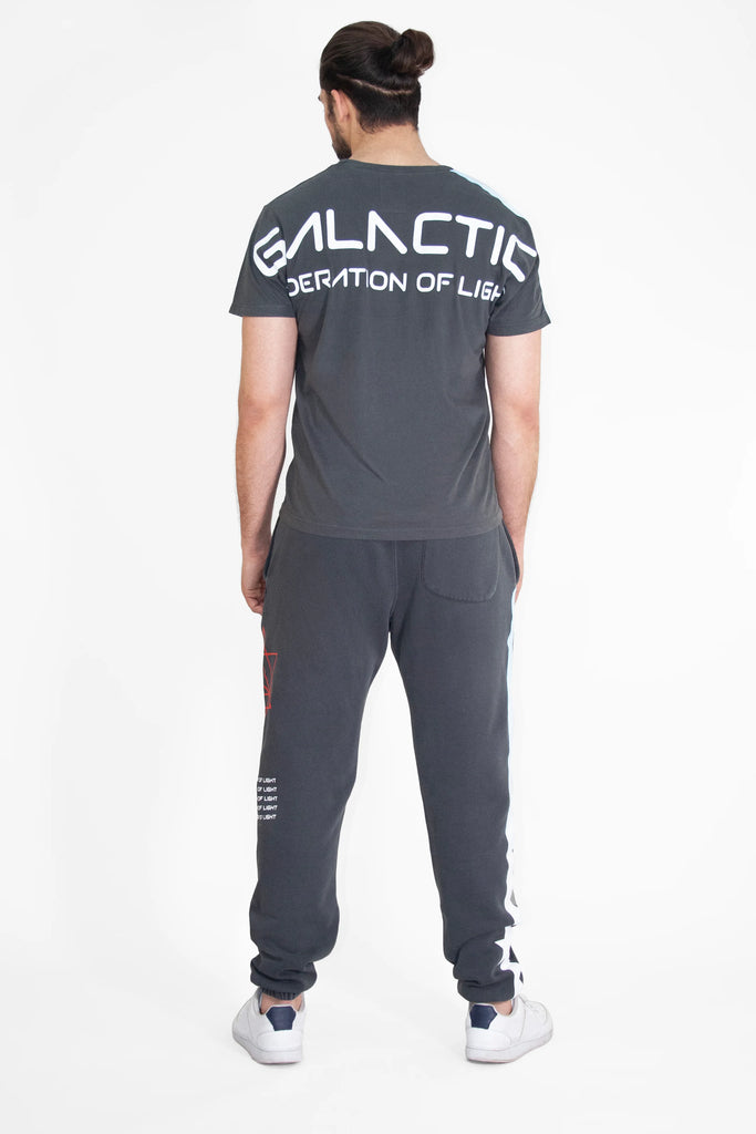 A person with a bun stands facing away, wearing a dark gray GFLApparel GFL Stars Tee in Space Glow with "GALACTIC FEDERATION OF LIGHT" written on the back. The relaxed fit t-shirt, made from breathable cotton, is paired perfectly with matching sweatpants.