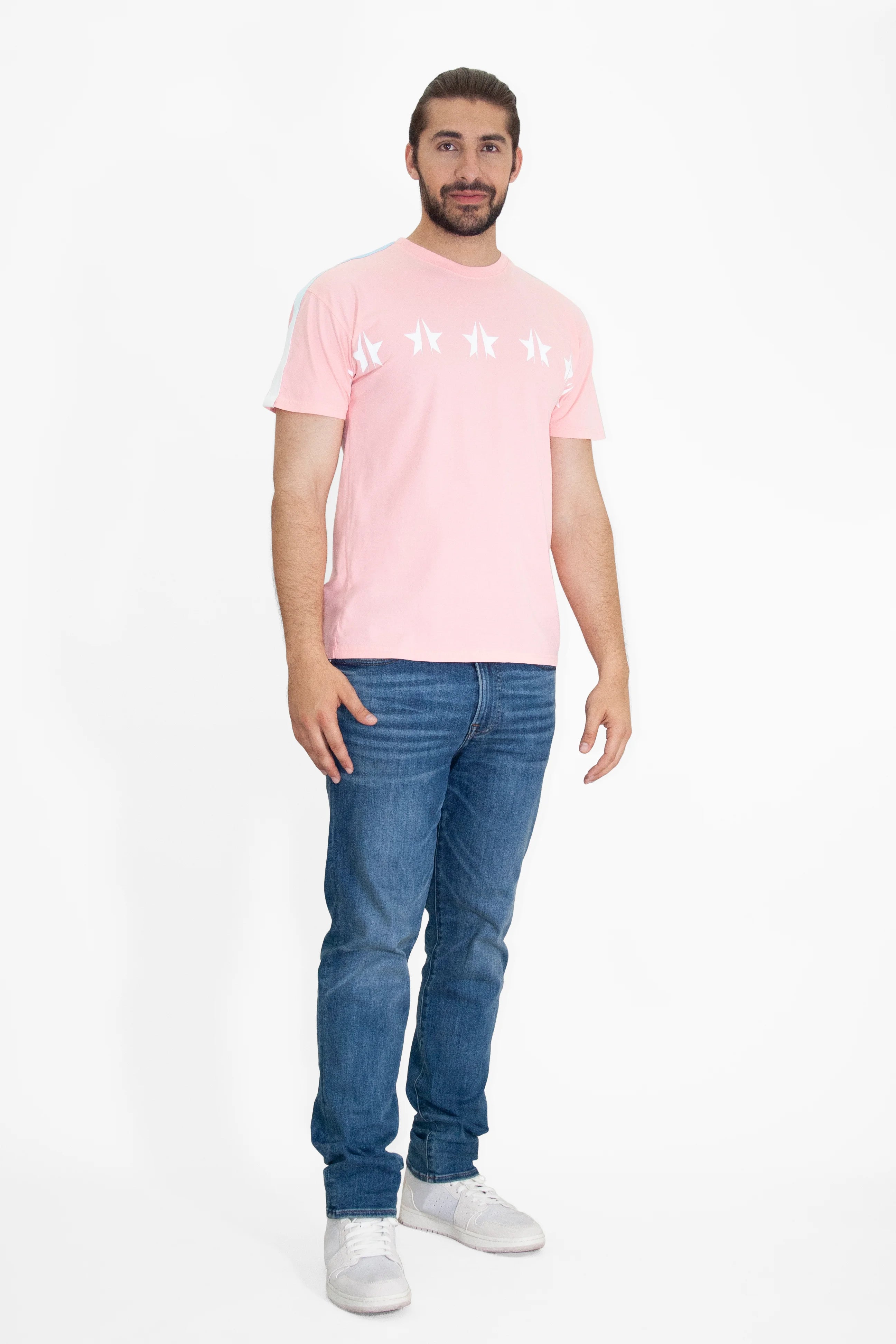 A man stands against a white background wearing a light pink GFLApparel GFL Stars Tee in Sunfade boasting white stars on the chest, paired with blue jeans and white sneakers. The relaxed fit of the breathable cotton T-shirt adds to its comfort and style.