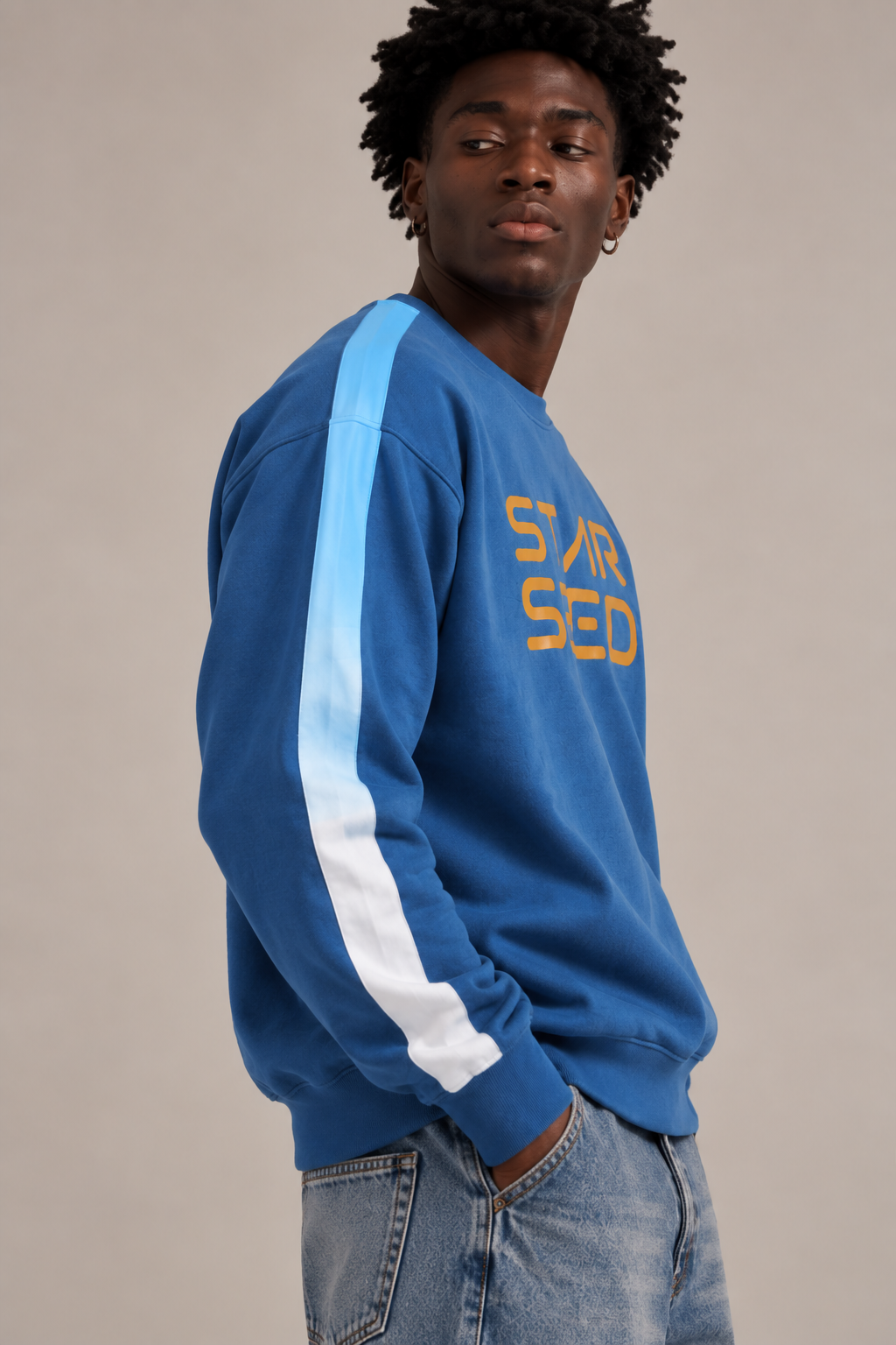 A man wearing a blue crewneck sweatshirt with a light blue to white stripe down the sleeve and yellow 'STAR SEED' text on the chest, paired with light-wash jeans, against a plain beige background.