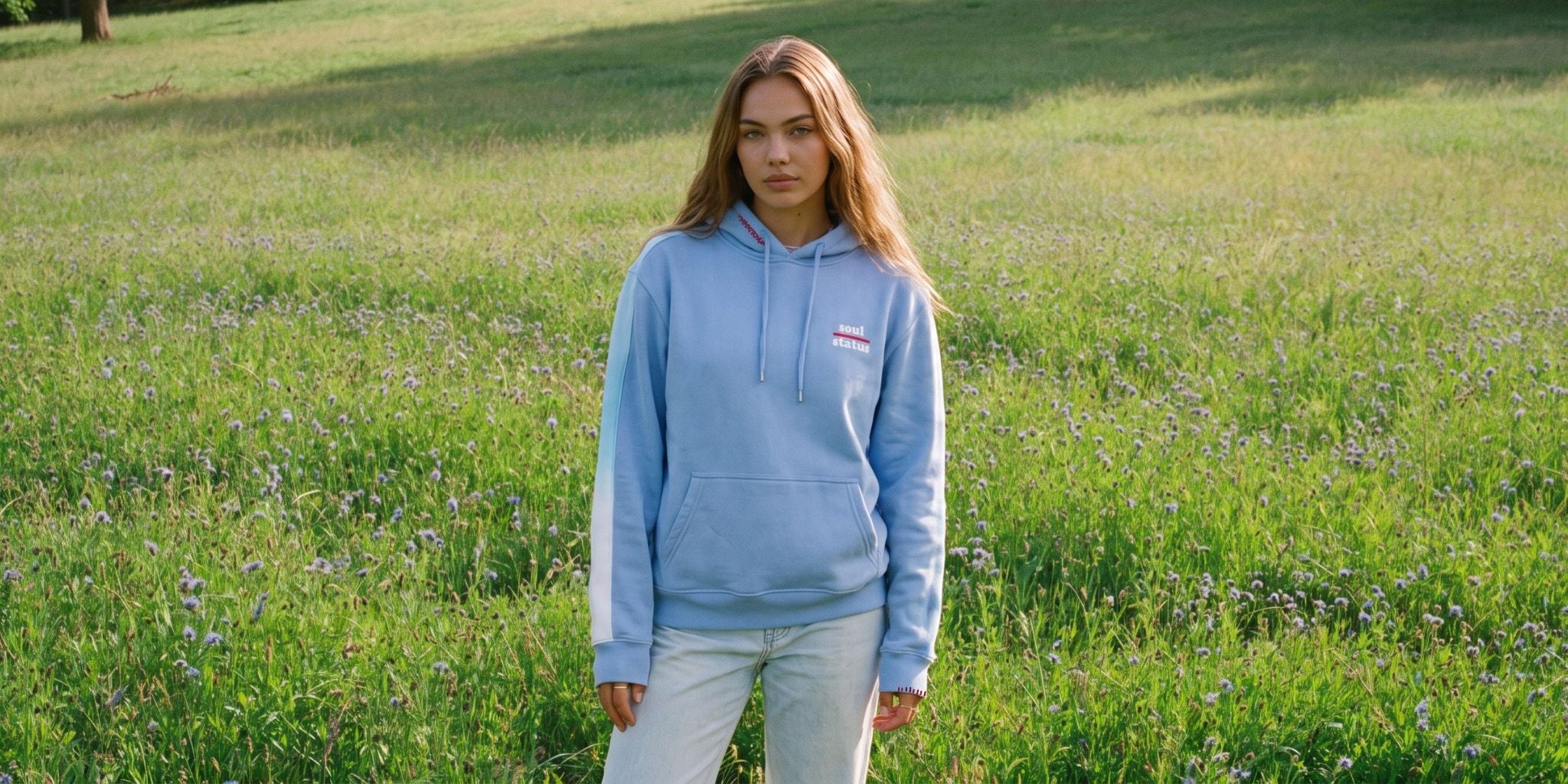 A woman standing in a grassy field wearing a light blue pullover hoodie with white sleeve stripes and a small red, white, and blue chest logo, paired with light gray pants.