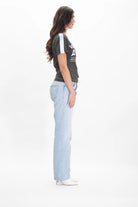 A woman with long hair stands in profile wearing a White Hat Army Tee in Caladan from GFLApparel, light blue jeans, and white heeled shoes. The background is plain white.
