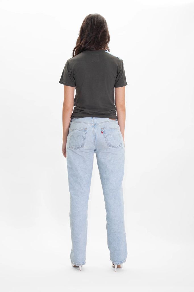 A person with long hair is standing with their back facing the camera, wearing a White Hat Army Tee in Caladan by GFLApparel and light blue jeans.