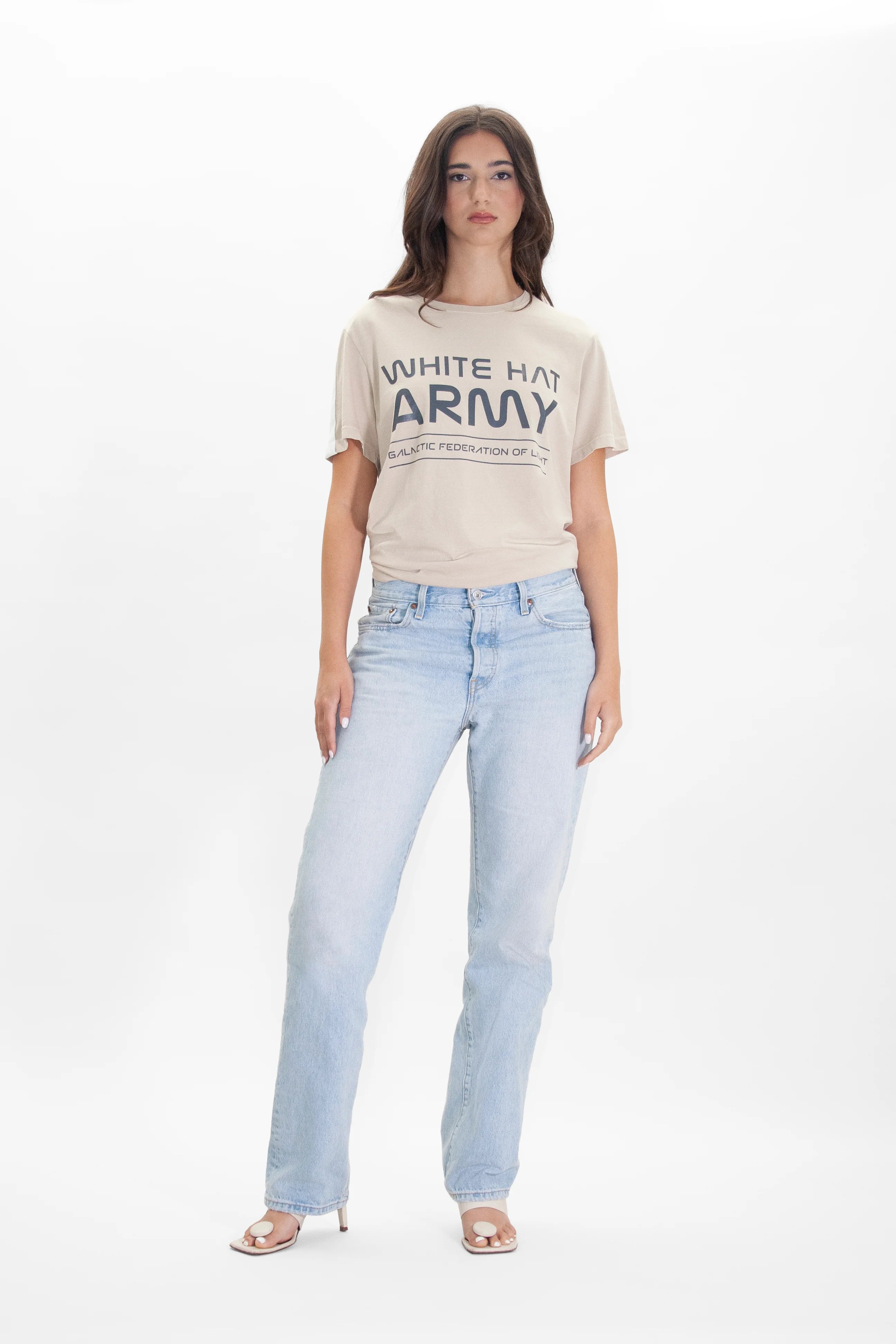 A woman stands against a plain background wearing a GFLApparel White Hat Army Tee in Dune and light blue jeans. She is also wearing open-toe heels.