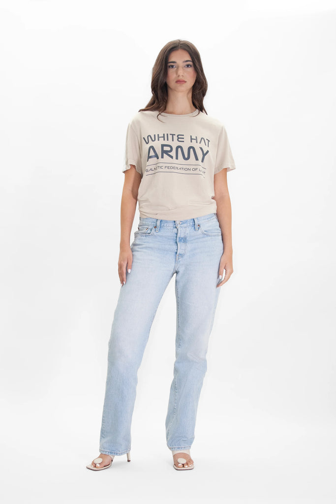 A woman stands against a plain background wearing a GFLApparel White Hat Army Tee in Dune and light blue jeans. She is also wearing open-toe heels.