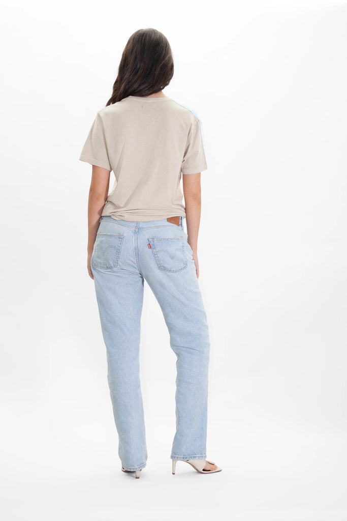 A person with long hair stands facing away, wearing a beige GFLApparel Illuminated Tee in Dune made from soft and breathable cotton and light blue jeans with a red tag on the back pocket, and high-heeled sandals, against a plain white background.