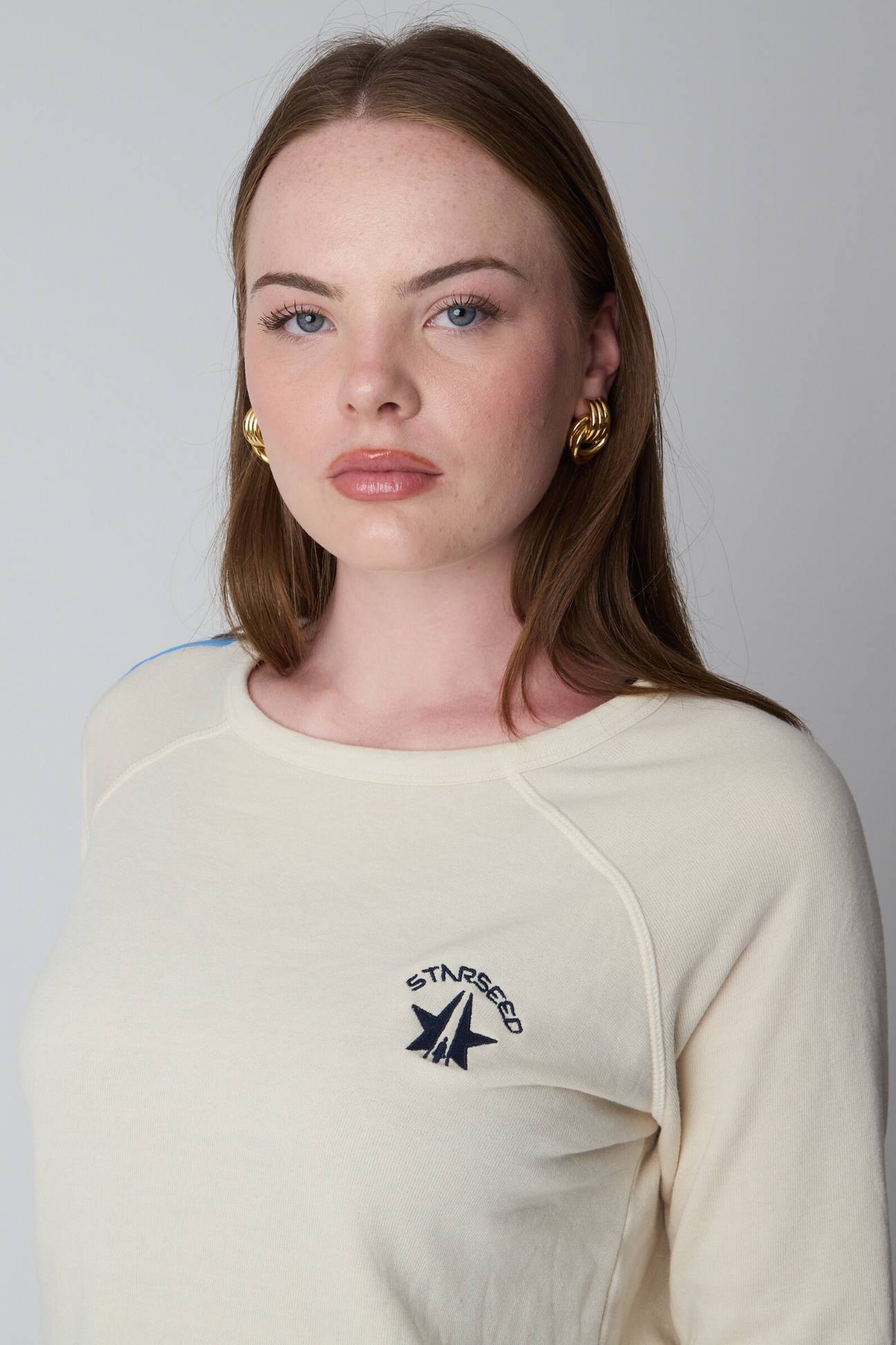 Woman wearing a cream-colored sweatshirt with a logo on a plain background