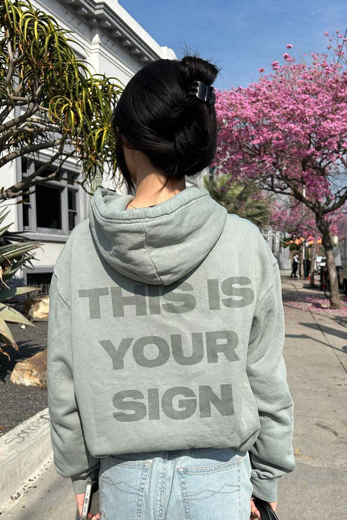 Woman in a gray, high-quality cotton-blend hoodie with the text "Angel Number 1111 Hoodie" from GFLApparel standing on a street lined with blooming pink trees.