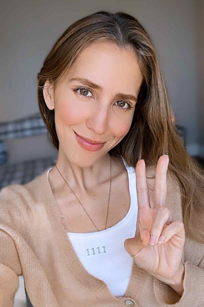 Woman in beige cardigan and white top smiling at the camera, making a peace sign with her right hand, embodying the positive energy of GFLApparel's Angel Number 1111 Cropped Tank in Bone.