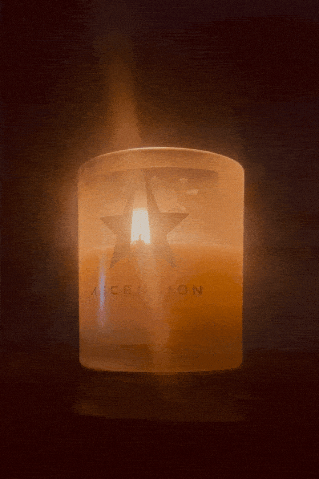 A lit Ascension Candle by Galactic Federation of Light in a frosted glass holder, adorned with a star and the word "ASCENSION," casts a warm glow in a dark setting. Made from non-carcinogenic soy wax infused with essential oils, it creates an ambiance that is both soothing and eco-friendly.