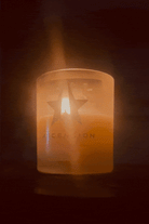 A lit Ascension Candle by Galactic Federation of Light in a frosted glass holder, adorned with a star and the word "ASCENSION," casts a warm glow in a dark setting. Made from non-carcinogenic soy wax infused with essential oils, it creates an ambiance that is both soothing and eco-friendly.