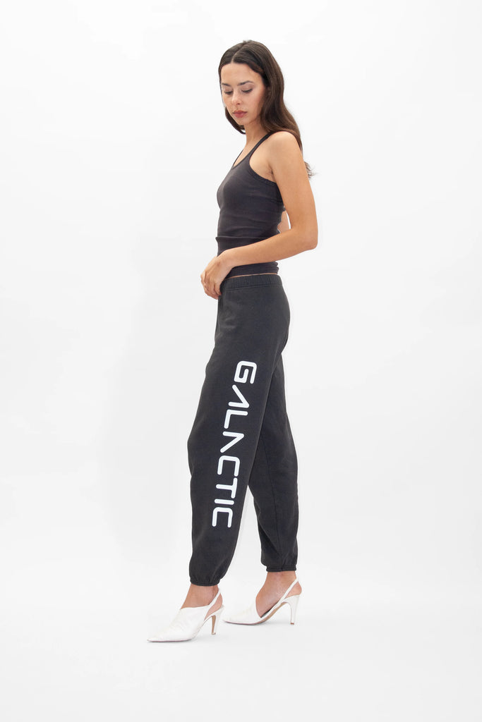 Woman in a black sleeveless top and GFLApparel Galactic Sweatpants in Space Glow with the word "GALACTIC" printed on the leg, standing in profile against a plain white background. She is wearing white high-heeled shoes.