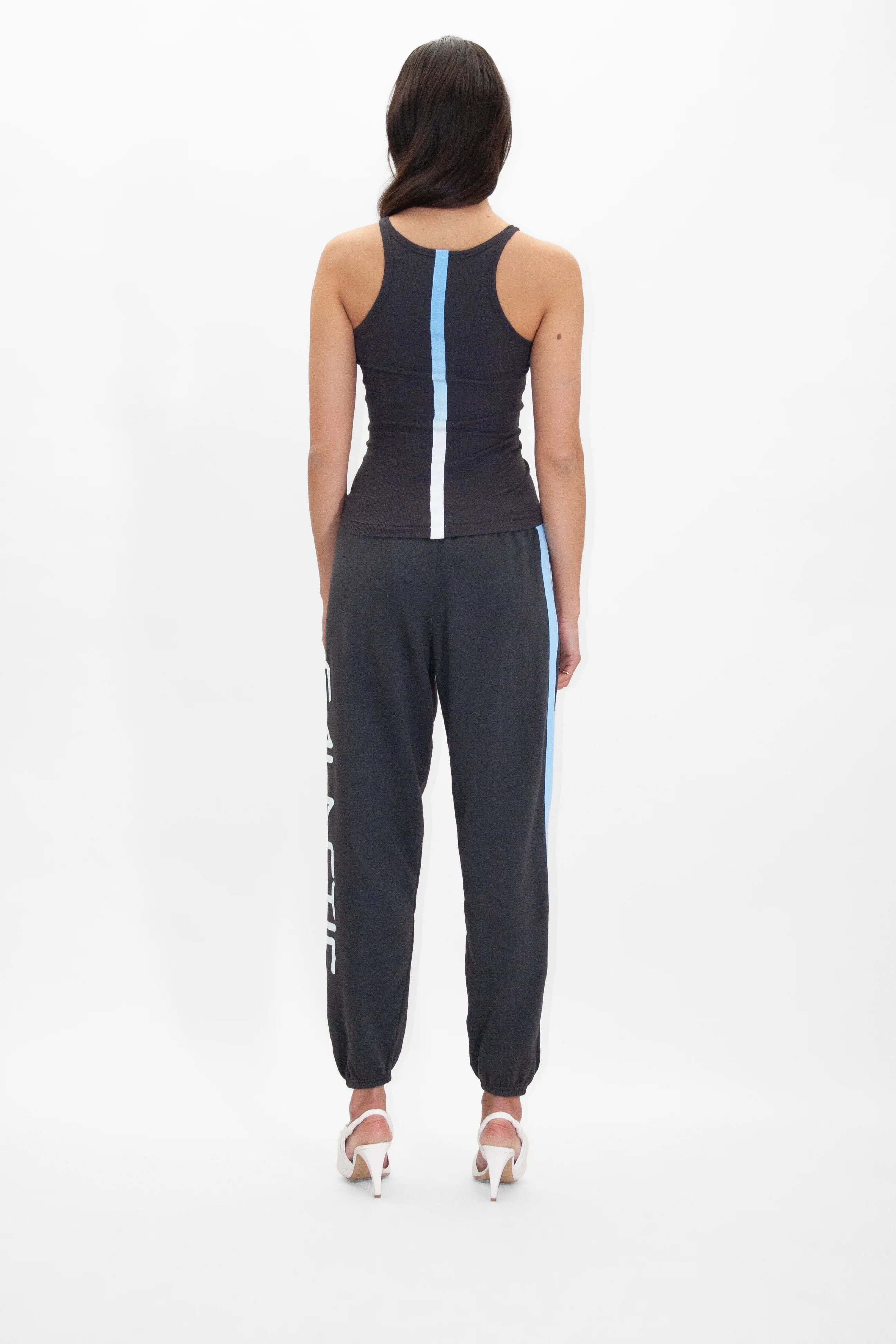 A person with long, dark hair stands facing away, wearing a black tank top with a white and blue vertical stripe at the back, Galactic Sweatpants in Space Glow from GFLApparel with a vertical stripe on one leg, and white high heels.