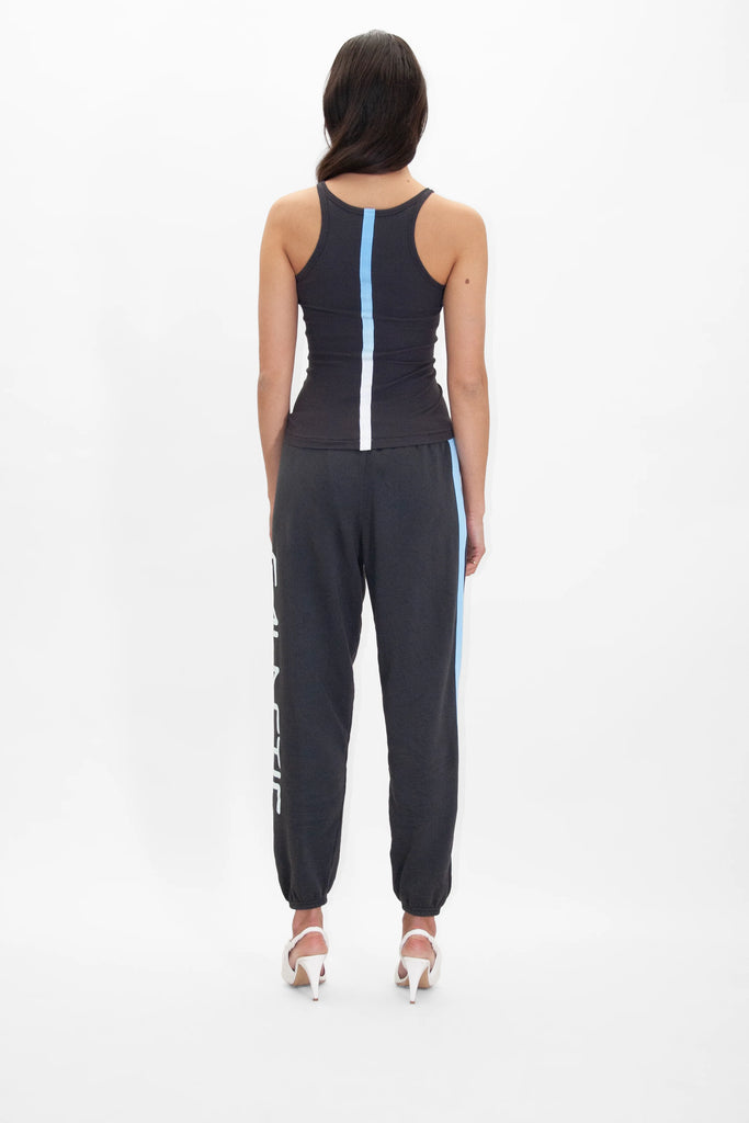 A person with long, dark hair stands facing away, wearing a black tank top with a white and blue vertical stripe at the back, Galactic Sweatpants in Space Glow from GFLApparel with a vertical stripe on one leg, and white high heels.