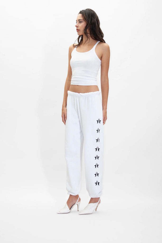 A person stands against a plain background wearing a white tank top, GFLApparel GFL Stars Sweatpants in Lite Beam adorned with black star symbols on the side, and white high heels.