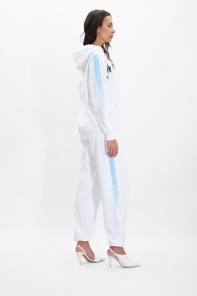 A woman stands in profile wearing a white hoodie and matching GFLApparel GFL Stars Sweatpants in Lite Beam with light blue stripes, paired with white high-heeled shoes, against a plain white background.