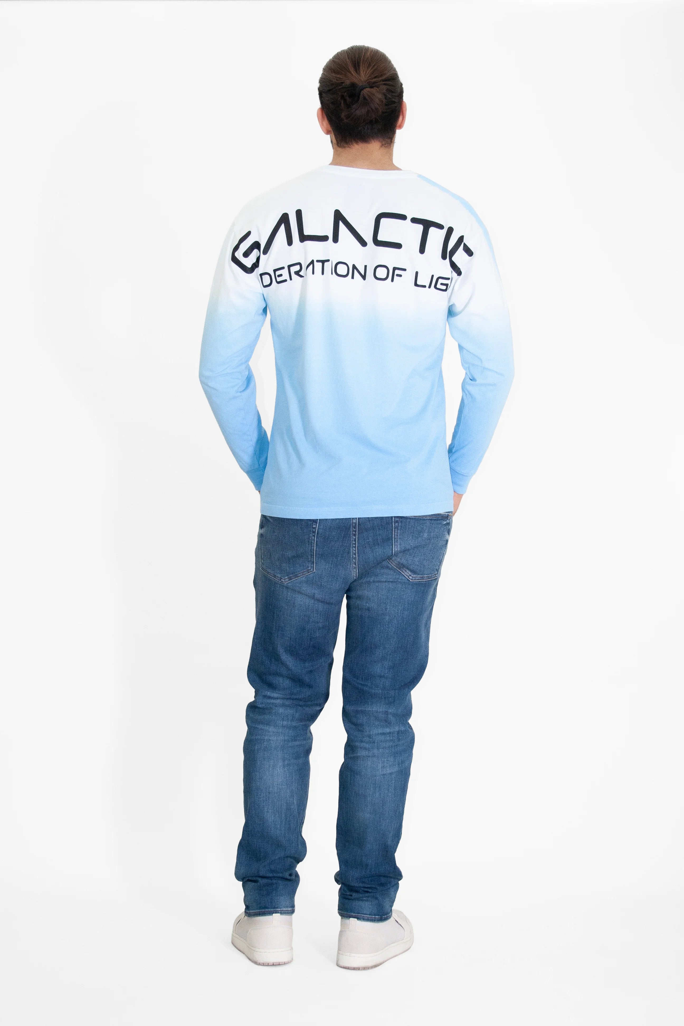 A person stands facing away from the camera, wearing jeans and a crewneck-style ombré long-sleeve shirt with 