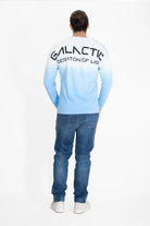 A person stands facing away from the camera, wearing jeans and a crewneck-style ombré long-sleeve shirt with "GFLApparel GFL Stars L/S in Atmosphere" printed on the back in large letters.