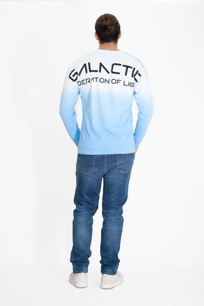 A person stands facing away from the camera, wearing jeans and a crewneck-style ombré long-sleeve shirt with "GFLApparel GFL Stars L/S in Atmosphere" printed on the back in large letters.