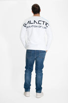 A person with long hair in a bun wears a white long-sleeve shirt, the **GFL Stars L/S in Lite Beam** by **GFLApparel**, paired with blue jeans and white shoes, standing against a white background. Notably, their attire hints at an affiliation with the GFL Stars.