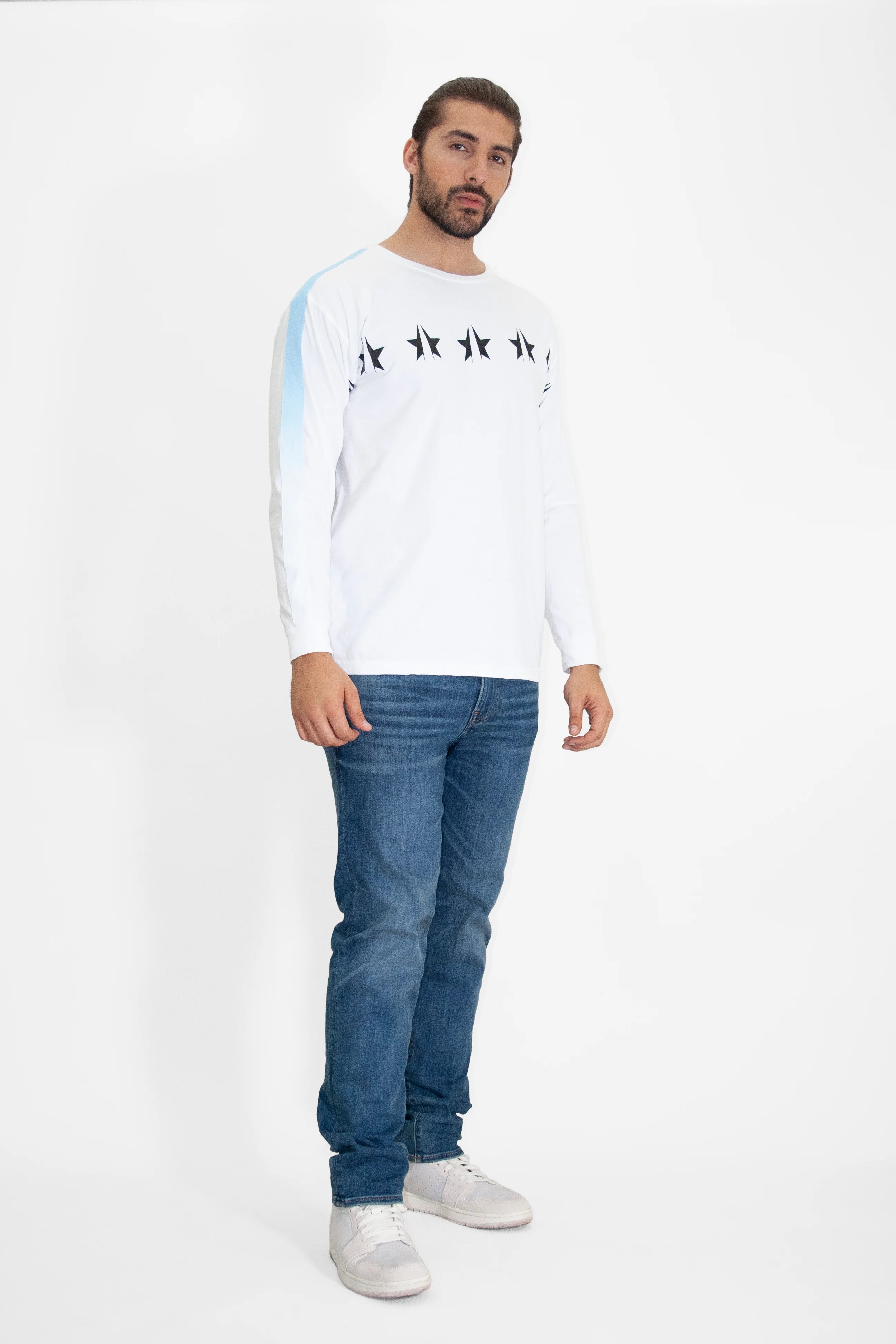 A man is standing against a plain background, wearing a white long-sleeved shirt with GFL stars patterns across the chest, blue jeans, and white sneakers. He has a neutral expression and is facing the camera. The shirt he is wearing is the GFL Stars L/S in Lite Beam from GFLApparel.