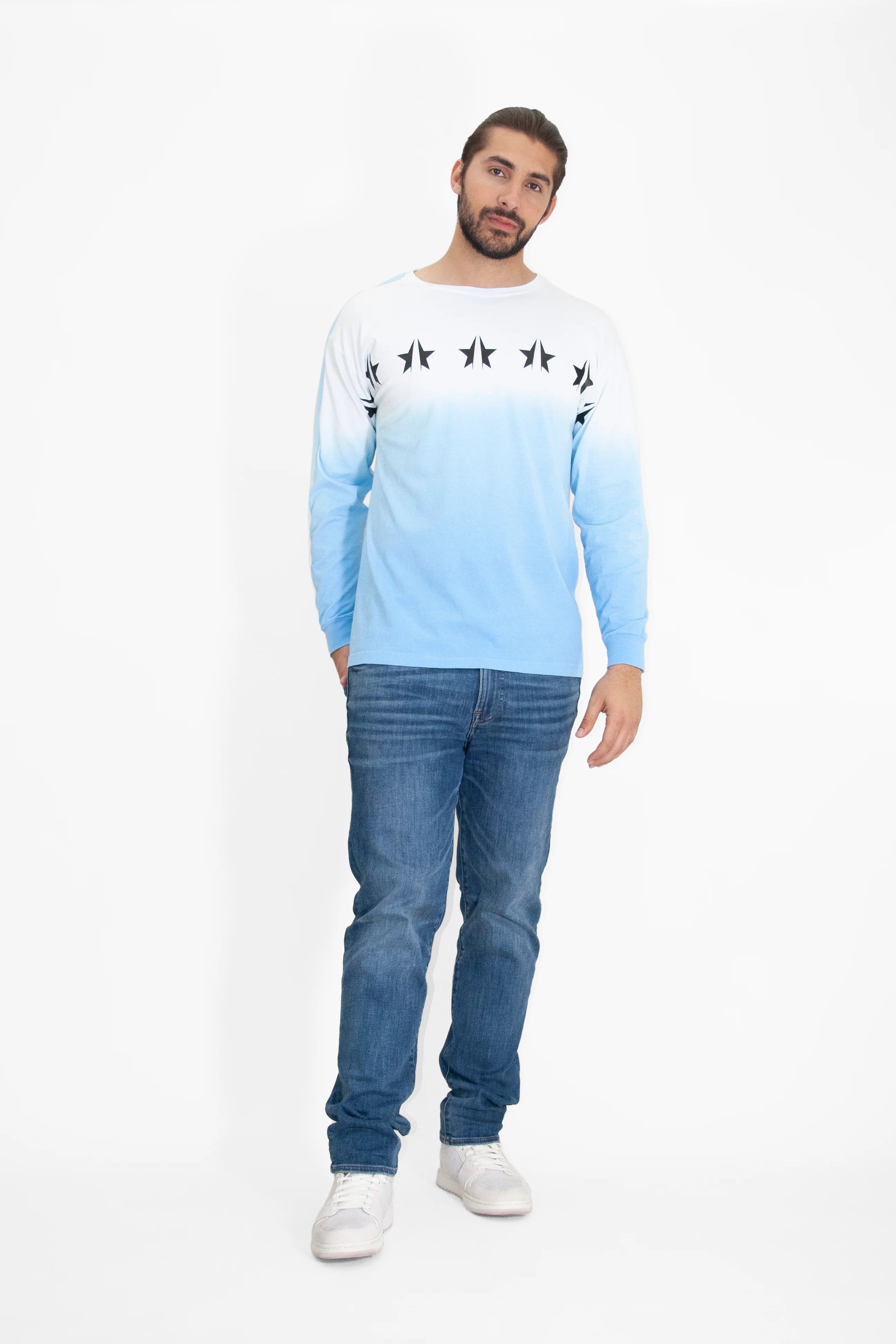 A bearded man stands against a white background wearing a crewneck-style GFLApparel GFL Stars L/S in Atmosphere, blue jeans, and white sneakers.