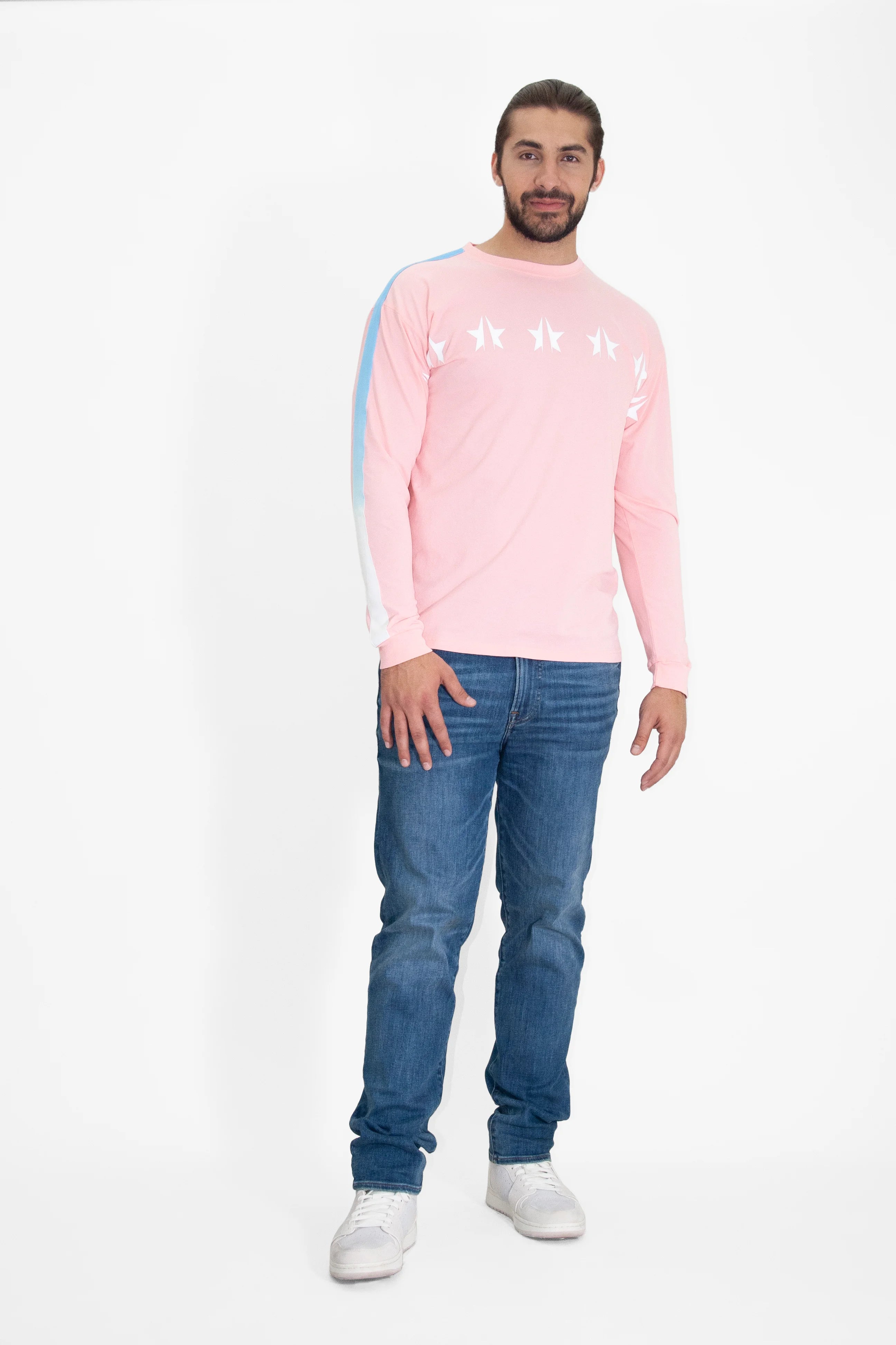 A man with long hair and a beard is wearing a relaxed fit, GFL Stars L/S in Sun Fade by GFLApparel, blue jeans, and white sneakers. He is standing against a plain white background.