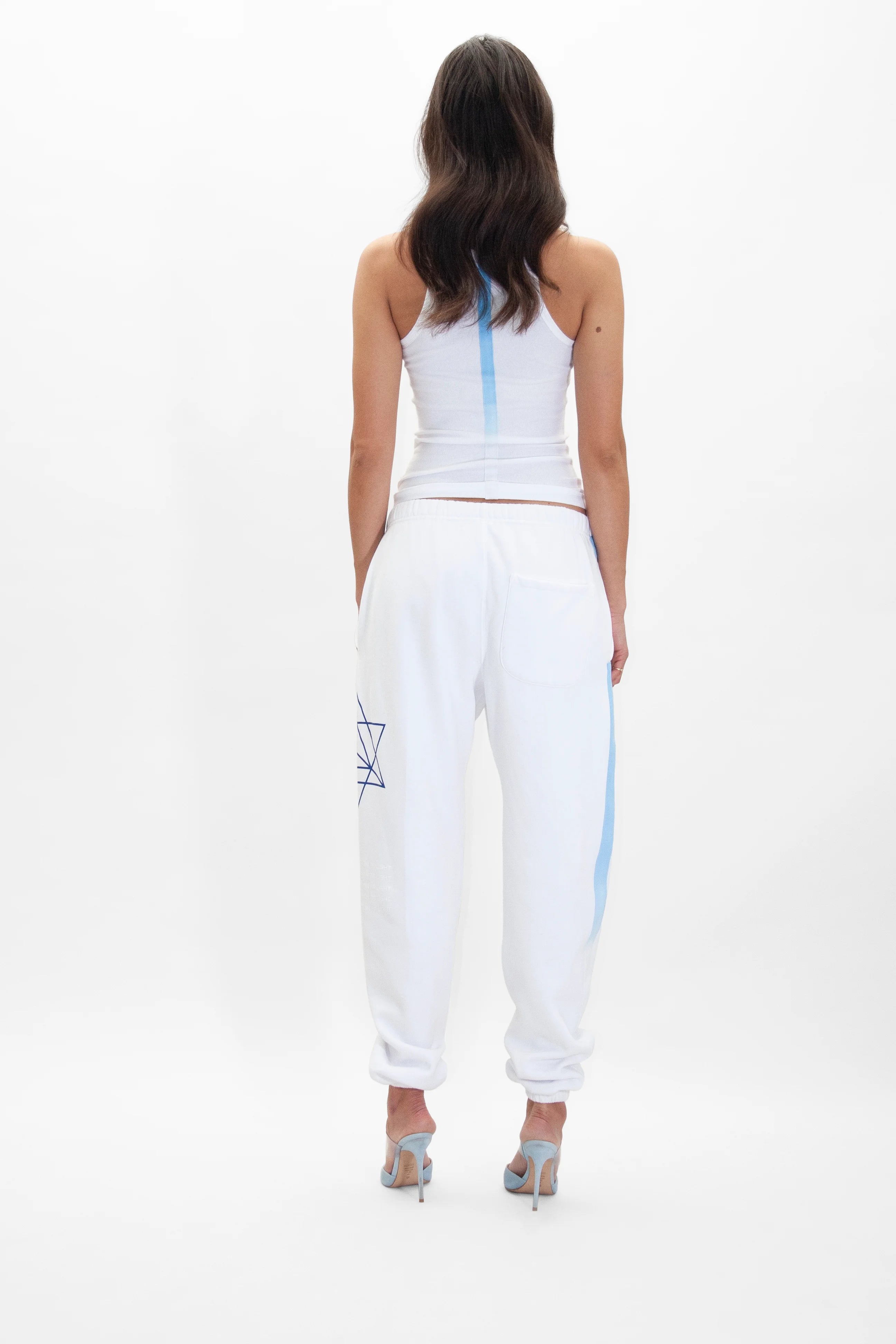 A person with long hair is standing facing away, wearing a white tank top, GFLApparel's Hypergalactic Pants in Lite Beam with a blue geometric design and a drawstring waistband, and light blue heels.