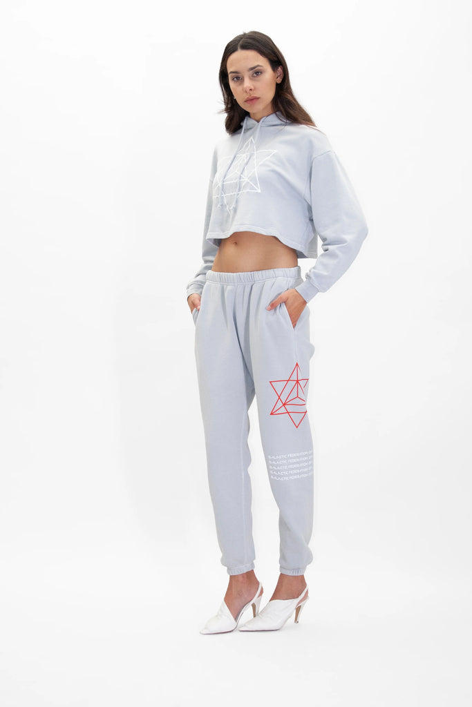 A woman in a light gray cropped hoodie and matching Hypergalactic Pants in Galactic Gray by GFLApparel with geometric designs. She is standing with hands in pockets, showcasing the drawstring waistband, and wearing white heeled shoes.