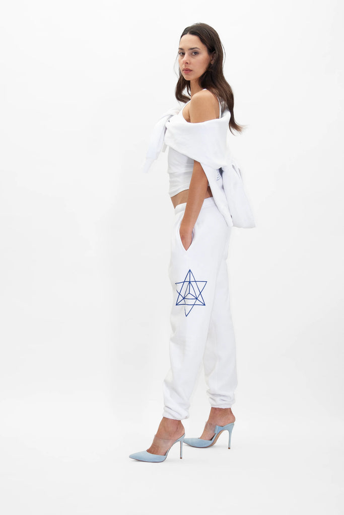 A person stands sideways with hands in pockets wearing a white off-shoulder top, GFLApparel Hypergalactic Pants in Lite Beam featuring a blue geometric design and a drawstring waistband, and blue high heels against a plain white background.