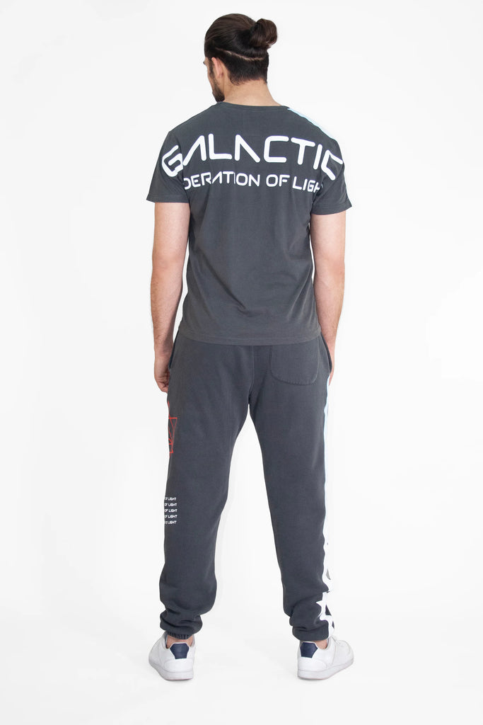 A person with dark hair in a bun is seen from behind, wearing a gray "Galactic Federation of Light" T-shirt and GFLApparel Hypergalactic Pants in Space Glow featuring a drawstring waistband, standing on a white background.