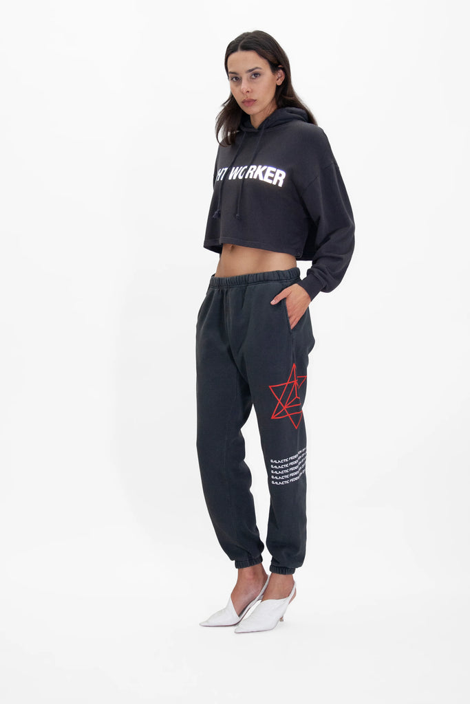 A woman stands against a white background wearing a cropped hoodie with "CITY WORKER" text, black Hypergalactic Pants in Space Glow by GFLApparel featuring a red geometric design and a drawstring waistband, paired with white pointed high heels.