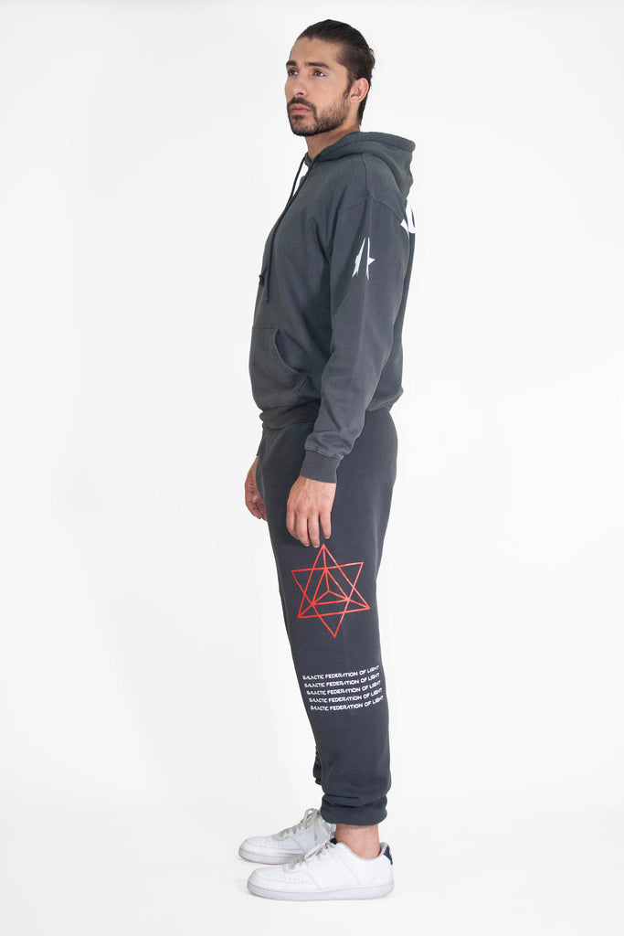 A person stands sideways, wearing a dark hoodie and GFLApparel Hypergalactic Pants in Space Glow with a red geometric star and white text on the left leg. The pants feature a drawstring waistband and are crafted from heavyweight cotton blend. They are also wearing white sneakers. The background is plain white.