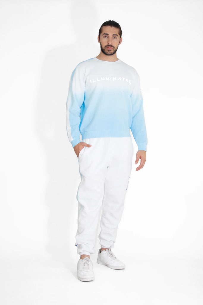 A man stands against a white background wearing an Illuminated Crewneck in Atmosphere by GFLApparel, paired with white pants and sneakers. He has short, dark hair and a beard.
