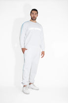 A man with a beard is standing against a white background, wearing a cozy Light Worker Crewneck in Lite Beam from GFLApparel with "Lightworker" in reflective print, white sweatpants, and white sneakers.