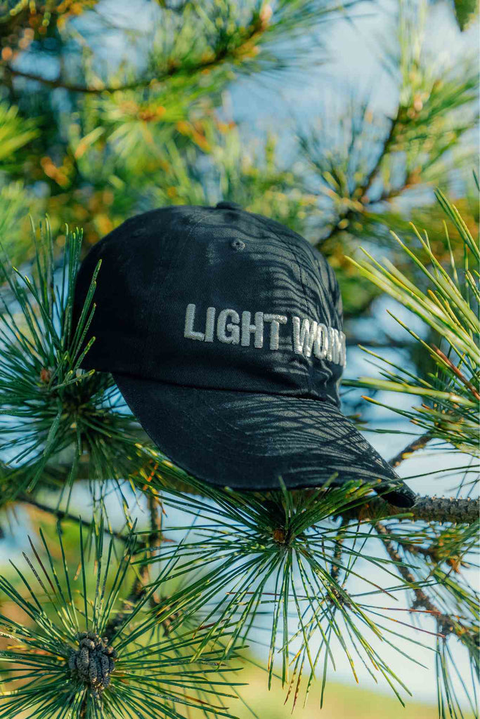 A black GFLApparel LIGHT WORKER BASEBALL CAP with the words "Light Worker" embroidered in white, hanging on a pine tree branch.
