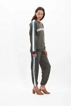 A woman stands wearing a green Light Worker Crewneck in Caladan by GFLApparel with the word "LIGHTWORK" in reflective print and matching green pants with light blue side stripes, both designed for a relaxed fit, paired with tan heeled sandals.