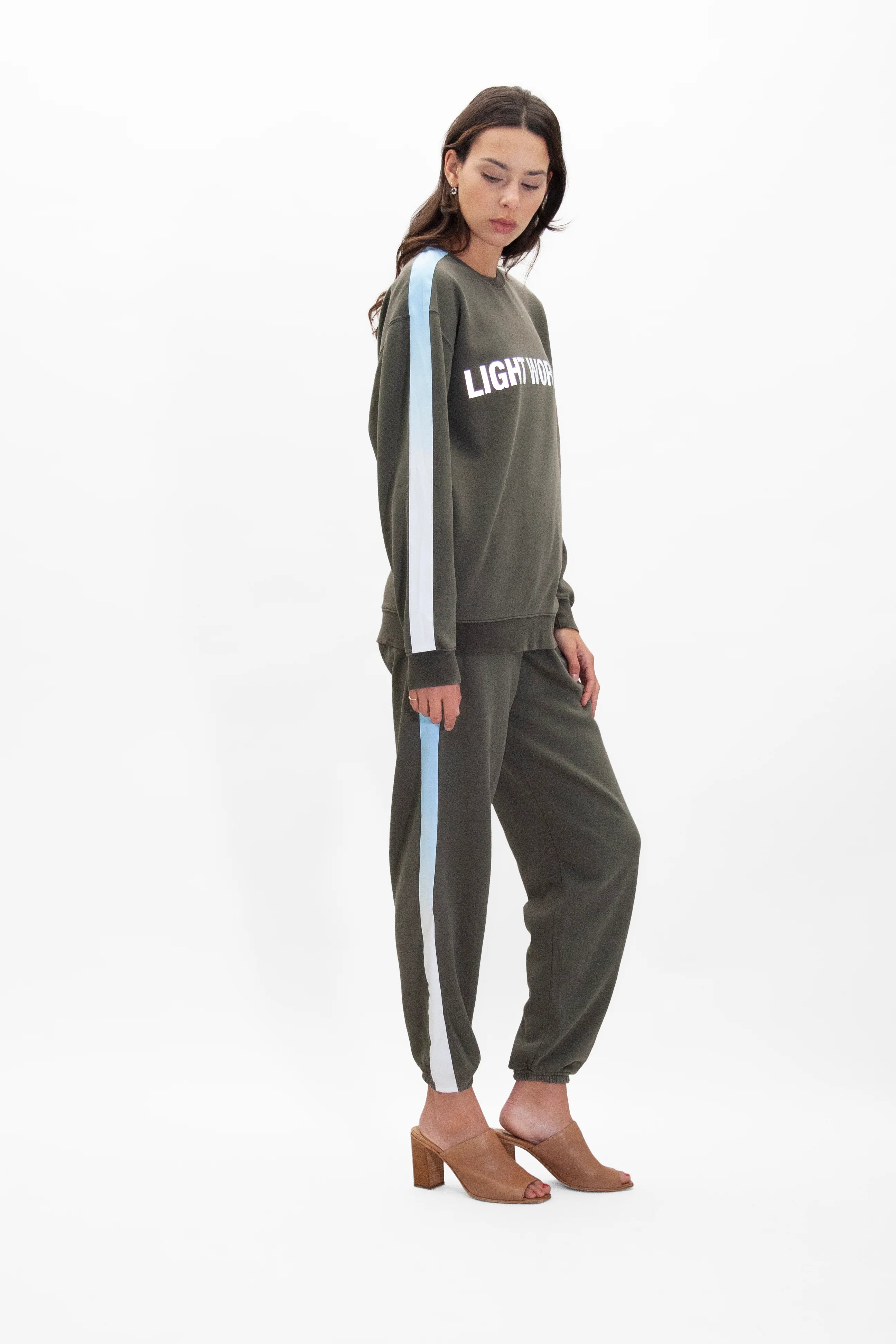 A woman stands wearing a green Light Worker Crewneck in Caladan by GFLApparel with the word "LIGHTWORK" in reflective print and matching green pants with light blue side stripes, both designed for a relaxed fit, paired with tan heeled sandals.