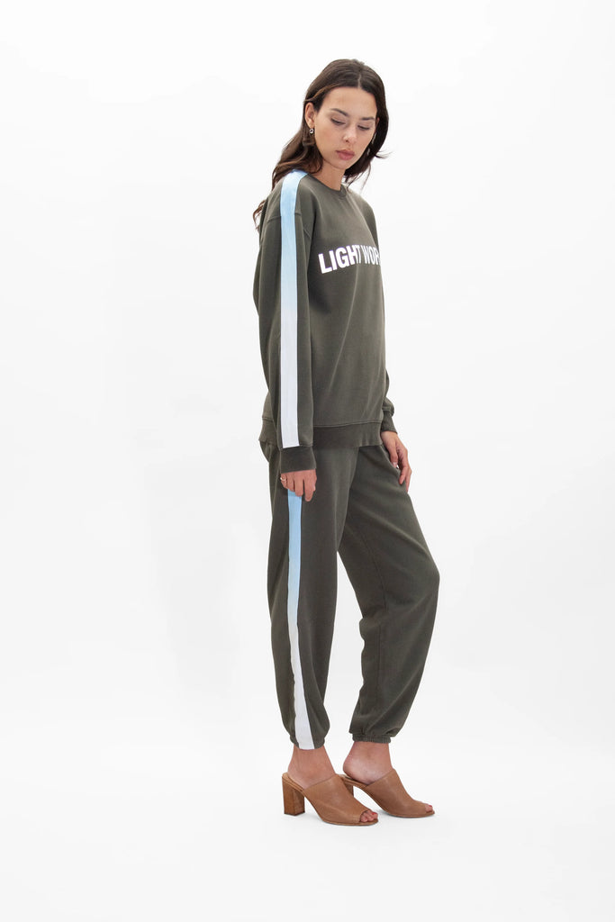 A woman stands wearing a green Light Worker Crewneck in Caladan by GFLApparel with the word "LIGHTWORK" in reflective print and matching green pants with light blue side stripes, both designed for a relaxed fit, paired with tan heeled sandals.