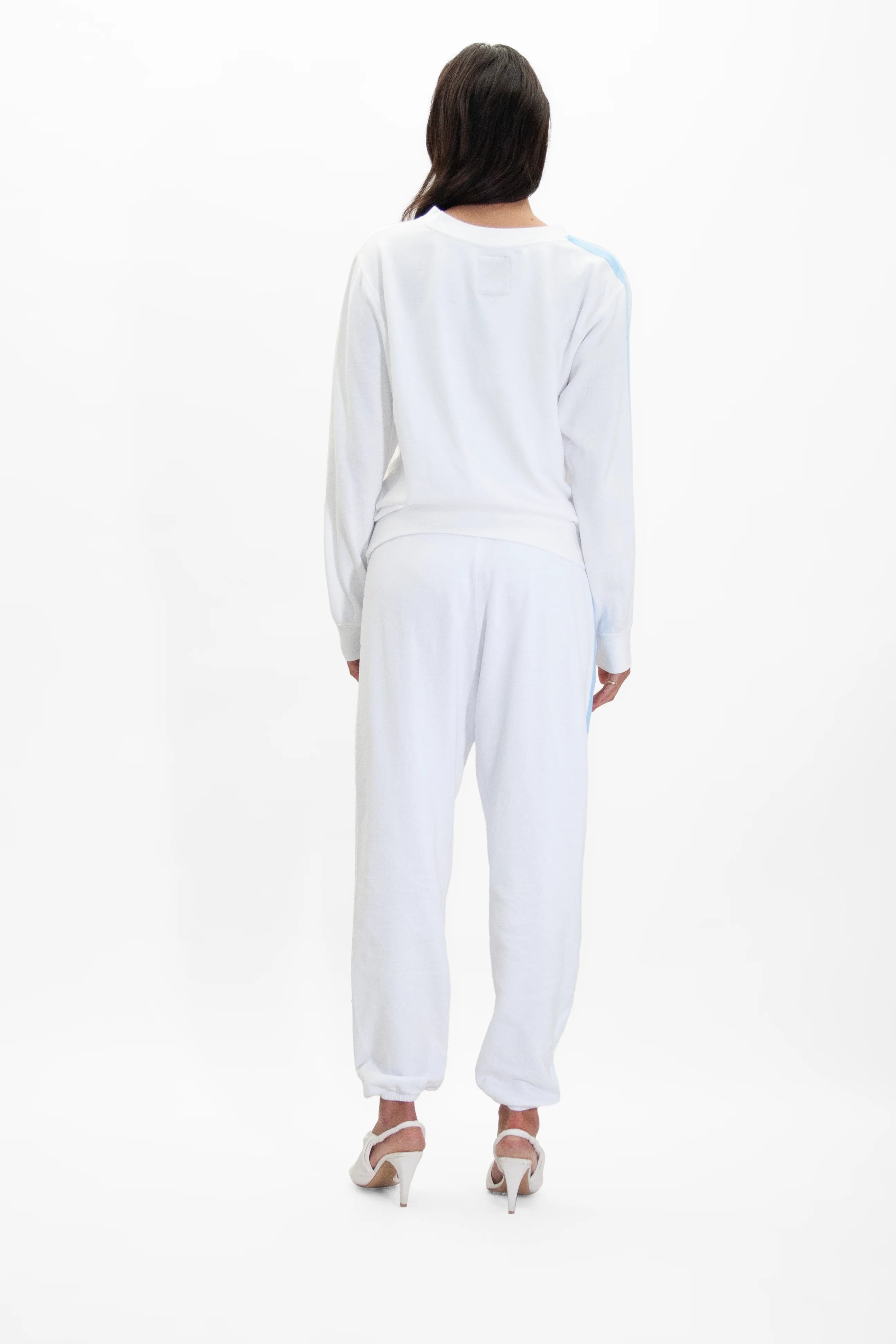 Woman standing and facing away from the camera, wearing a white GFLApparel Light Worker Crewneck in Lite Beam with reflective print and long sleeves and ankle-length pants. She has on white high-heeled shoes.
