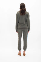 A person stands facing away from the camera wearing a green cotton Light Worker Crewneck in Caladan and matching sweatpants in a relaxed fit, and tan heels, with wavy brown hair cascading down their back. This stylish look is brought to you by GFLApparel.
