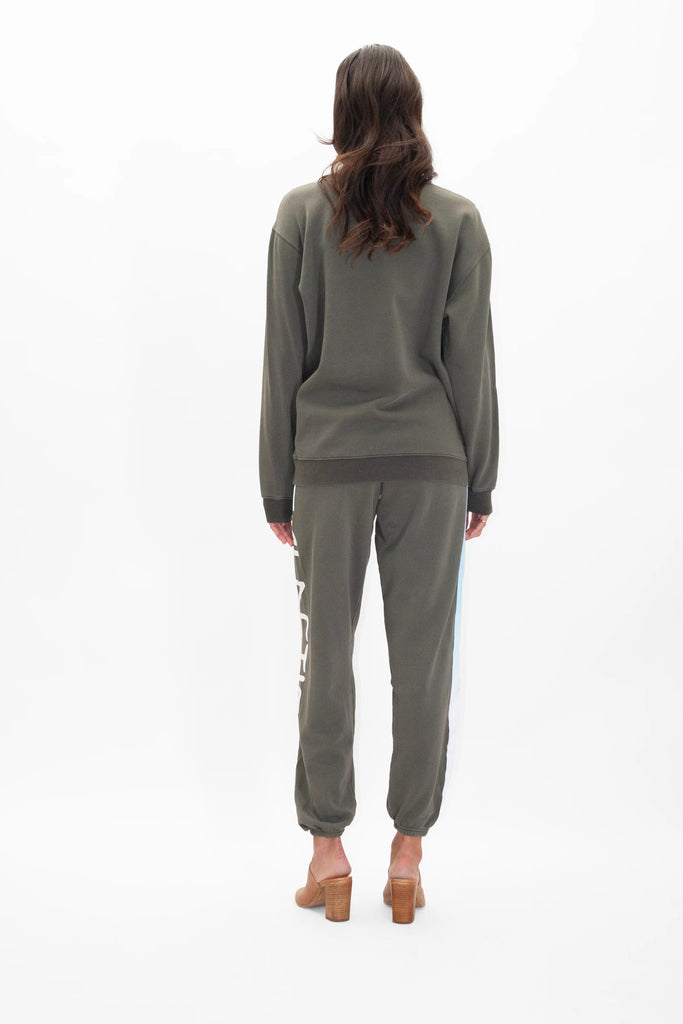 A person stands facing away from the camera wearing a green cotton Light Worker Crewneck in Caladan and matching sweatpants in a relaxed fit, and tan heels, with wavy brown hair cascading down their back. This stylish look is brought to you by GFLApparel.