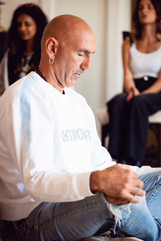 A bald man wearing a cozy GFLApparel Light Worker Crewneck in Lite Beam and jeans is sitting cross-legged indoors, engaged in a focused activity. Two women are sitting in the background.