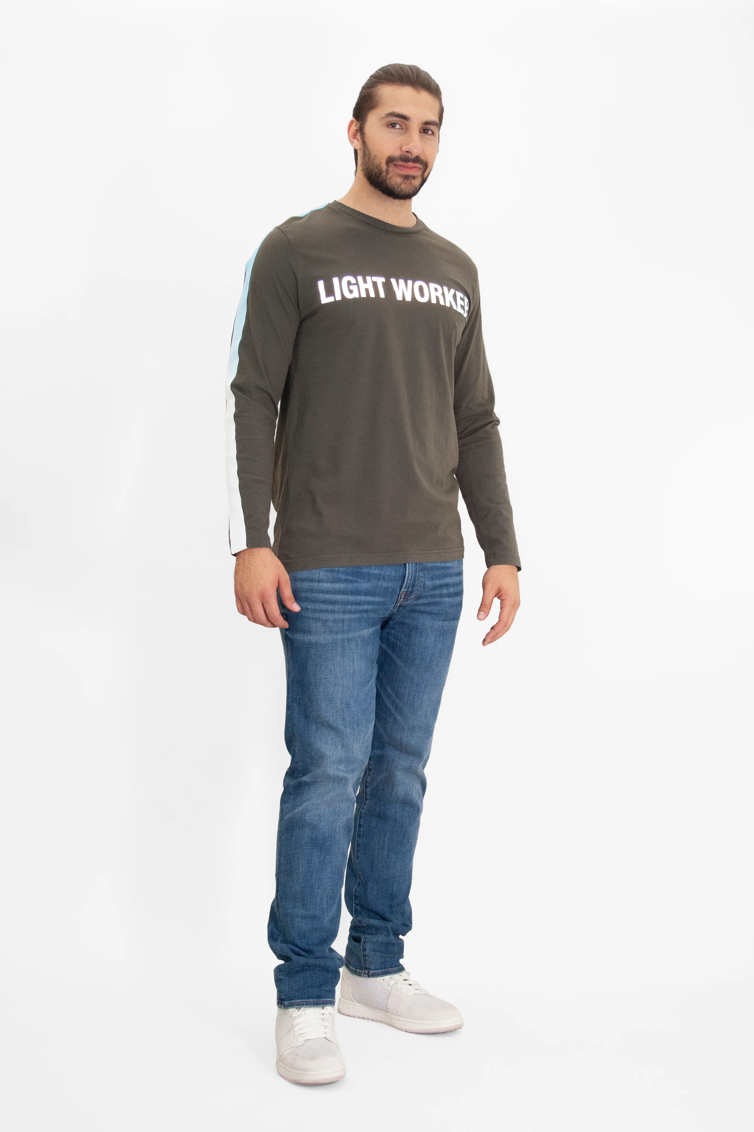 A man with long hair tied back stands against a white background, wearing a dark long-sleeve cotton T-shirt with "LIGHT WORKER" text in reflective print, blue jeans, and white sneakers. He is wearing the GFLApparel Light Worker L/S in Caladan.