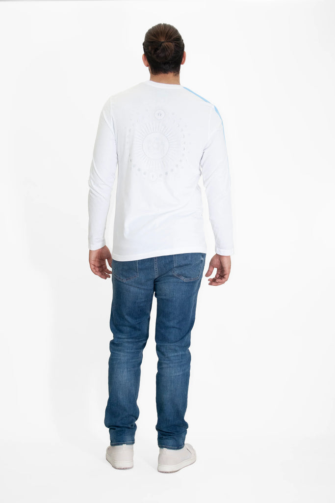 A person with long hair in a bun stands facing away, wearing a white long-sleeve cotton T-shirt from GFLApparel's Light Worker L/S in Lite Beam collection, blue jeans, and white sneakers. The background is plain white.