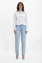 Woman with long dark hair wearing a white long-sleeve cotton T-shirt reading "Light Worker L/S in Lite Beam" in reflective print and light blue jeans, standing against a plain white background.