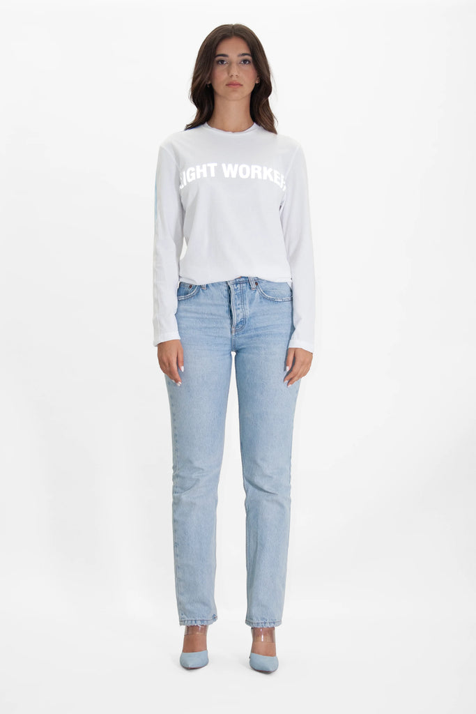 Woman with long dark hair wearing a white long-sleeve cotton T-shirt reading "Light Worker L/S in Lite Beam" in reflective print and light blue jeans, standing against a plain white background.