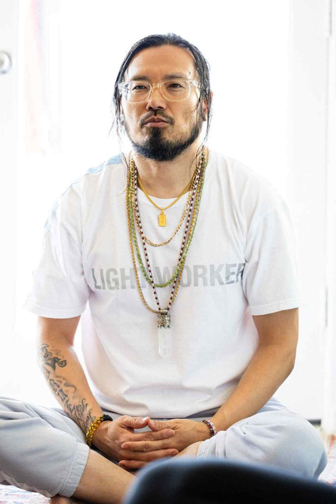 A person with long hair and glasses sits cross-legged wearing a white GFLApparel Light Worker Tee in Lite Beam made from soft breathable cotton, multiple beaded necklaces, and light-colored pants.