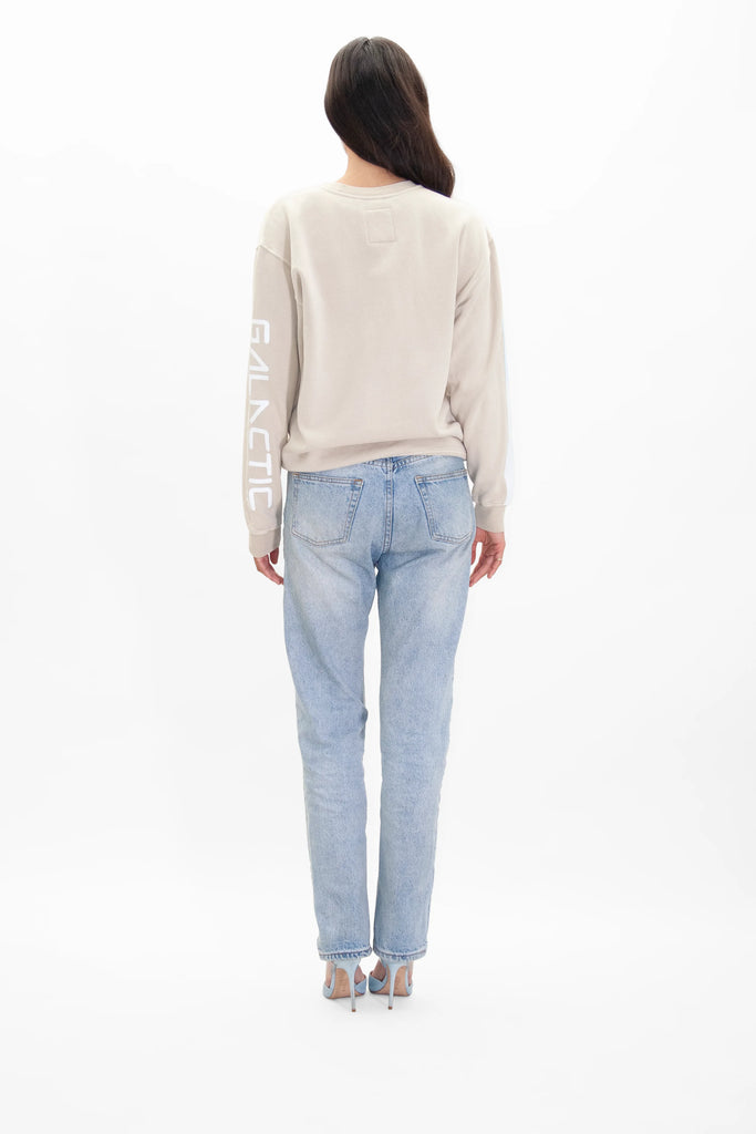 A person with long hair is standing with their back to the camera. They are wearing a beige sweatshirt with "GALACTIC" on the sleeve, light blue jeans, and high-heeled shoes. The sweatshirt, resembling a Merkaba Crewneck in Dune by GFLApparel, subtly hints at Kabbalah mysticism and Sacred Geometry themes.